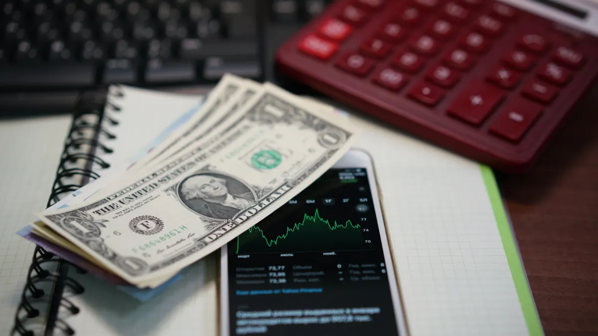 Dollar bills on a smartphone showing a stock price chart, next to a notebook and a red calculator