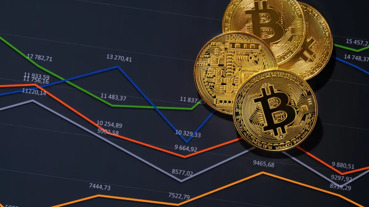 Four gold bitcoin coins resting on a dark screen showing colored price lines and numbers