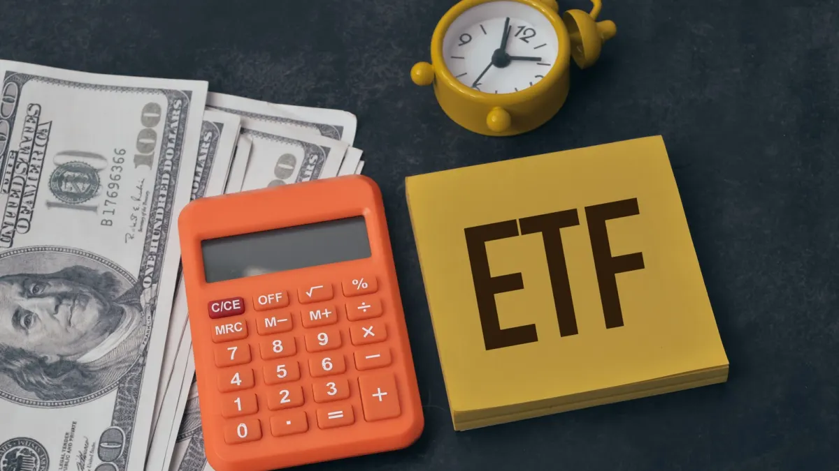 Yellow sticky note reading ETF next to an orange calculator, US hundred-dollar bills and a yellow alarm clock
