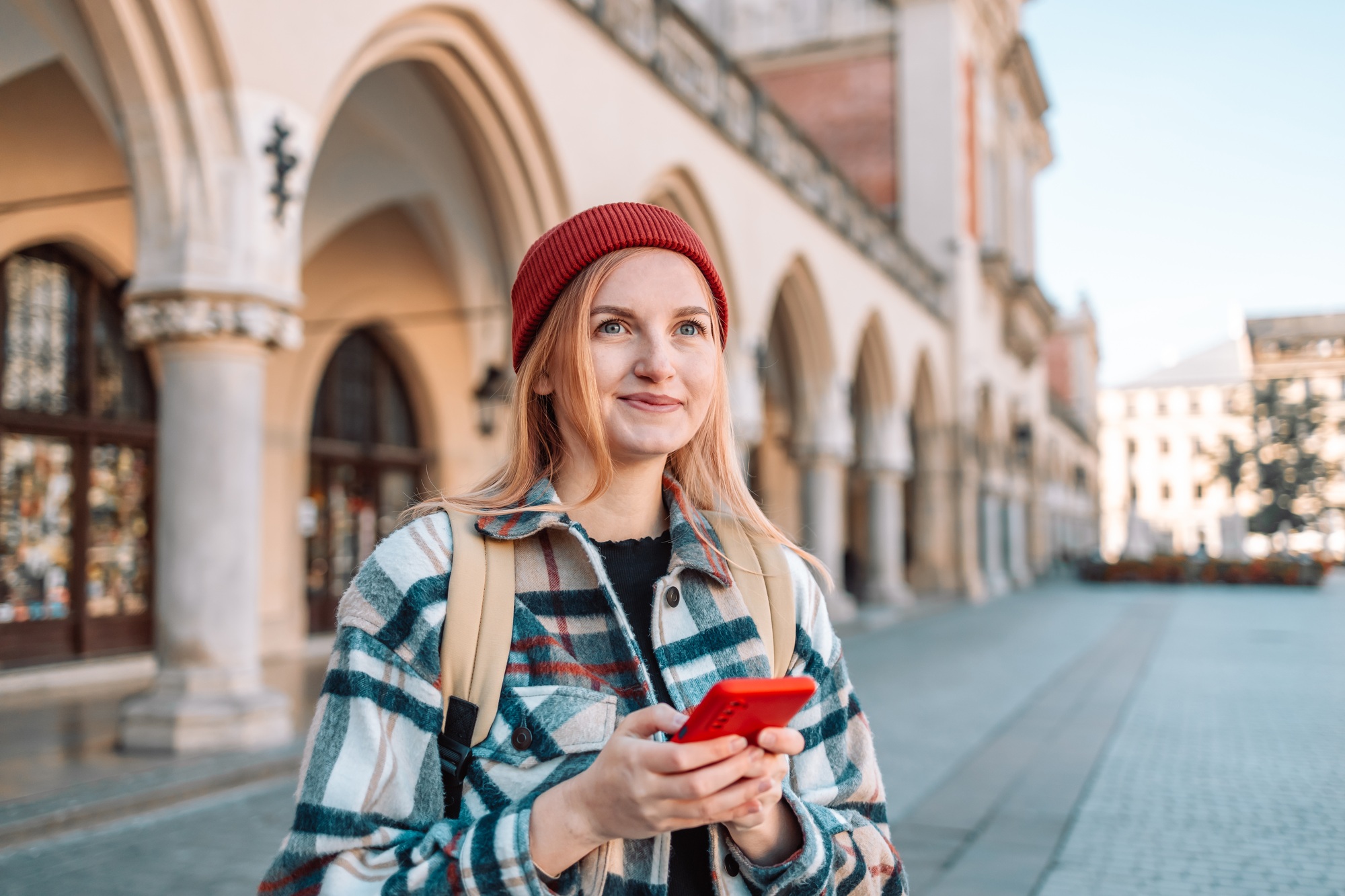 Happy hipster woman uses smartphone outdoors in big old city background.