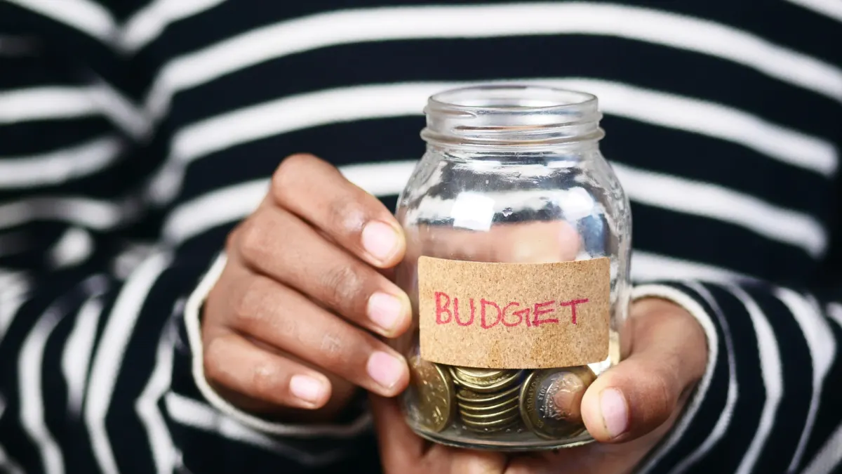 Hands holding a small glass jar of coins labeled Budget against a striped sweater