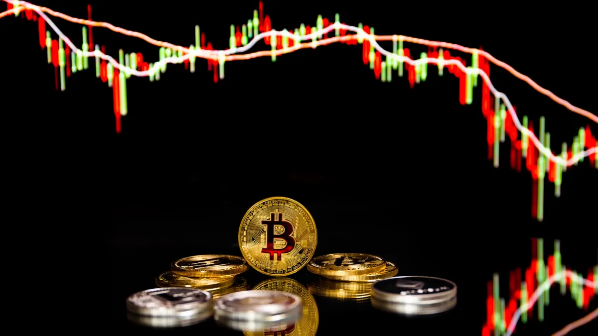 Gold and silver bitcoin coins on a reflective black surface beneath a red and green candlestick price chart