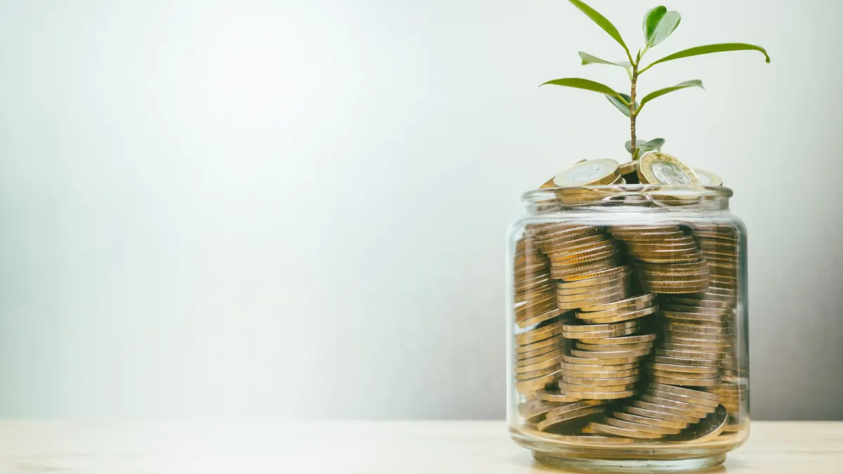 Glass jar filled with coins with a small green plant growing from the top, symbolizing growing savings